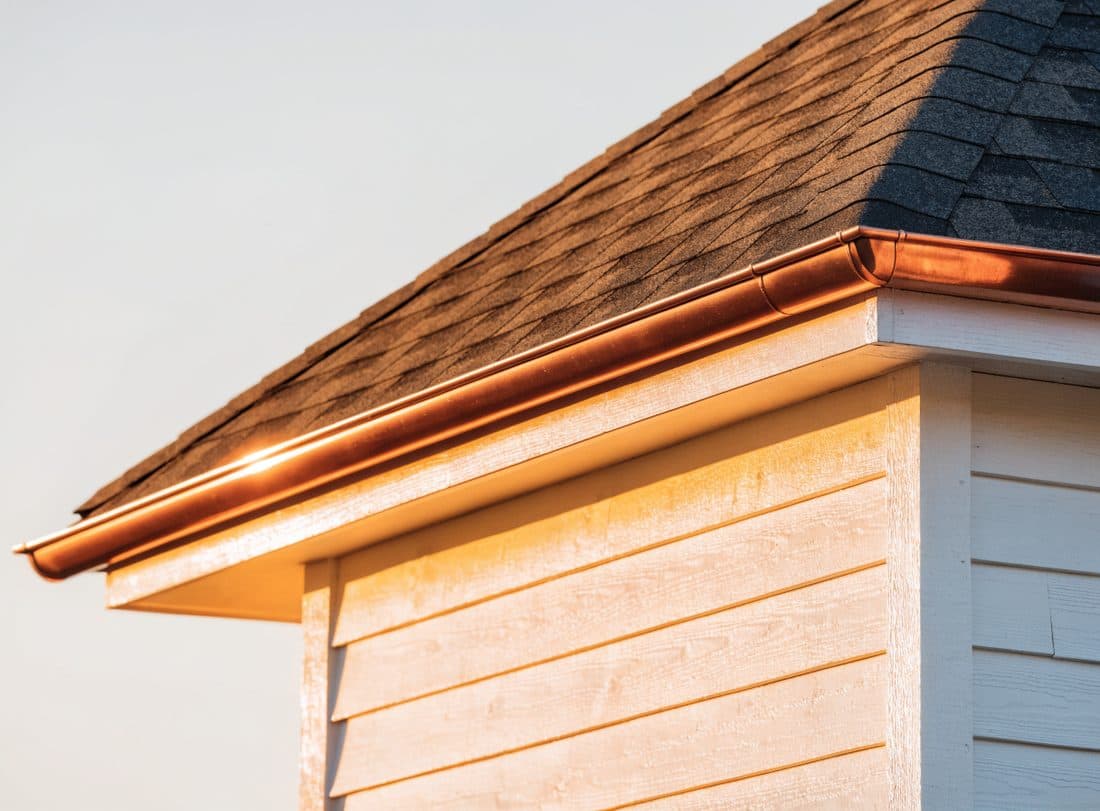The corner of a newly finished roof, charcoal shingles meeting a white fascia and a copper gutter against a blue sky.